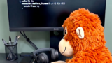 Orange plush monkey typing on a keyboard at a desk, with a monitor showing lines of code in the background